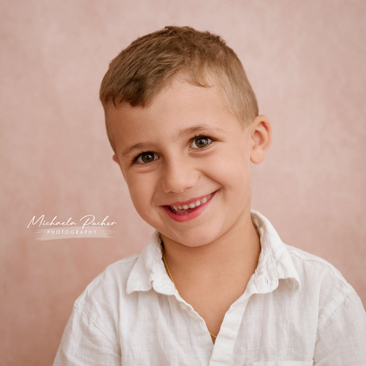 Lächelnder Junge mit kurz geschnittenem Haar, der in einem weißen Hemd vor einem sanften rosigen Hintergrund in die Kamera blickt.